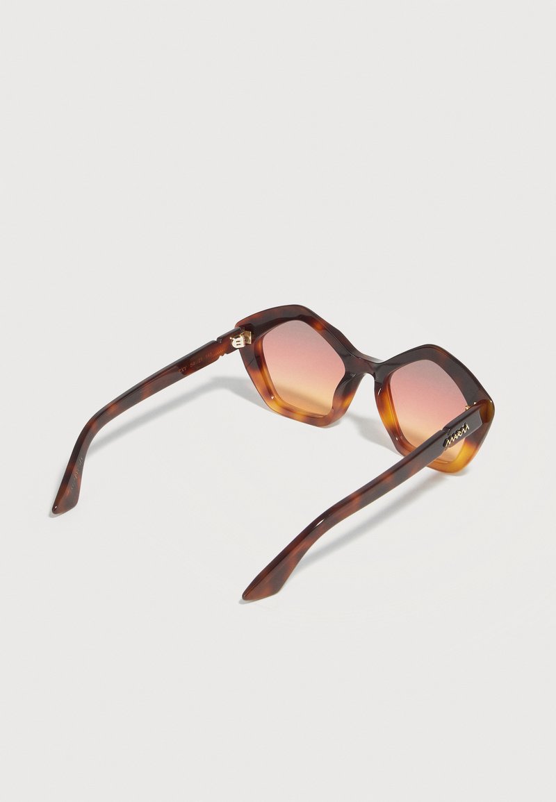 Lunettes de soleil carrées oversize en écaille de tortue avec des verres dégradés marron-orange et un détail doré de marque sur les branches, sur un fond uni.