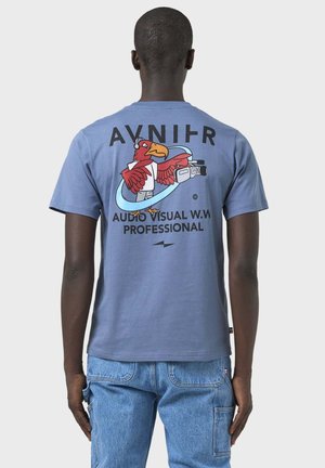 Person trägt ein blaues T-Shirt mit einem roten Vogel, der eine Videokamera hält, und dem Text „AVNIHR AUDIO VISUAL W.W PROFESSIONAL“ auf dem Rücken.