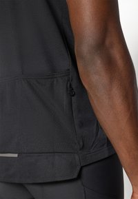 Schwarzes Sportshirt aus strukturiertem Stoff, mit einer Seitentasche und einem dezenten reflektierenden Akzent am Saum. Nahaufnahme des sichtbaren Arms.