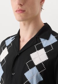Chemise noire à manches courtes avec un motif en losange bleu et blanc, fabriquée en tissu léger, avec un col pointu et une fermeture à boutons.