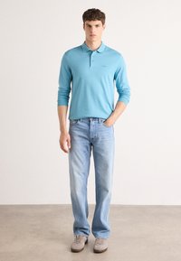 Svetlo modra polo majica z dolgimi rokavi in tremi gumbi, v kombinaciji s svetlim denim jeansom in sivimi čevlji, stoječa na nevtralni ozadju.