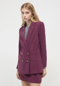 Giovane donna che indossa un blazer doppiopetto in velluto a coste viola e una gonna abbinata, in piedi contro uno sfondo bianco uniforme.