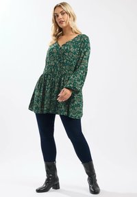 T tunica floreale verde con scollatura a V, maniche lunghe e orlo arricciato, abbinata a jeans skinny blu scuro e stivaletti neri con tacco.