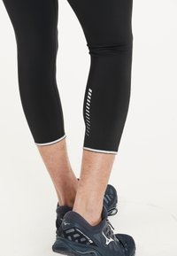 Svart atletiska leggings med reflexdetaljer, kortad design och vit randdetalj nära vaden. Mönstrad footwear kompletterar outfiten.