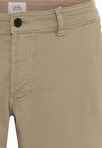 Pantalon beige décontracté avec passants de ceinture, fermeture par bouton noir, poches avant et étiquette de marque Camel Active à l’intérieur de la ceinture.