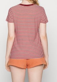 Kobieta w czerwono-białą paskowaną koszulę z krótkim rękawem i pomarańczowe szorty, pokazana z tyłu na jednolitym tle.