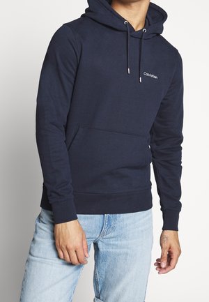 Mann trägt eine marineblaue Calvin Klein Kapuzenjacke mit Fronttasche und hellblaue Jeans, steht vor einem schlichten Hintergrund.