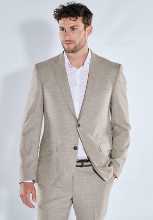 Mann mit lockigem Haar, der einen beigen Anzug und ein weißes Hemd trägt, steht mit einer Hand in der Tasche vor einem schlichten hellen Hintergrund.