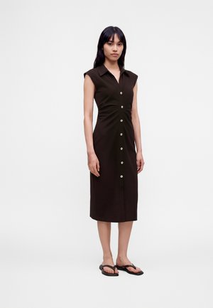 Frau, die frontal steht, trägt ein ärmelloses schwarzes midi-Kleid mit Knopfleiste und schwarze Sandalen mit offenen Zehen vor einem weißen Hintergrund.