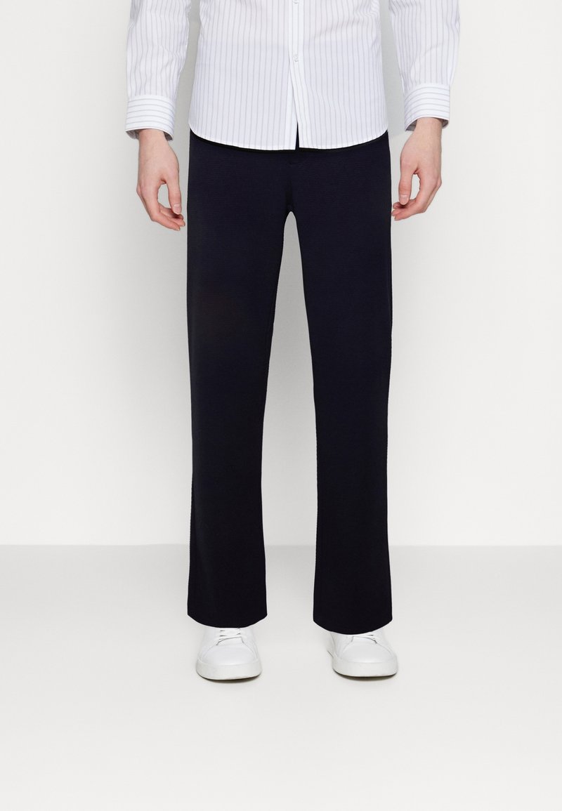 Navy blue pants in a straight-cut design. Made of a smooth fabric, they feature an elastic waistband and are paired with white sneakers.