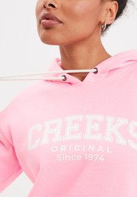 Felpa rosa con cappuccio dotata di un laccio con occhielli metallici, con la scritta "CREEKS ORIGINAL Since 1974" stampata in bianco sulla parte anteriore. Tessuto morbido al tatto.