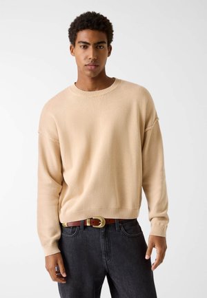 Beiger Strickpullover mit rundem Ausschnitt und überschnittenen Schultern, kombiniert mit dunkler Jeans und einem braunen Gürtel mit goldenem Verschluss.