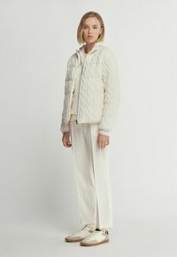 Vit puffjacka med quiltat mönster, som har en dragkedja fram och en huva, kombinerad med krämfärgade byxor och sneakers med beige accenter.