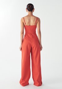 Žena s účesom do drdolu, oblečená v bezrukávovom širokom overalovom kostýme v hrdzavo-oranžovej farbe, stojí s chrbtom obráteným k divákovi na bielom pozadí.