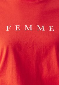 T-shirt en coton rouge avec le mot "FEMME" en lettres majuscules blanches, design col rond, texture douce et surface lisse.