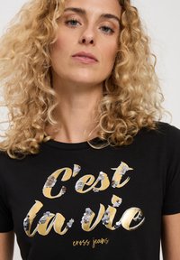 Czarna koszulka z krótkim rękawem i okrągłym dekoltem, ozdobiona złotymi i srebrnymi cekinami z napisem "C'est la vie" oraz "cross jeans". Miękka tekstura materiału.