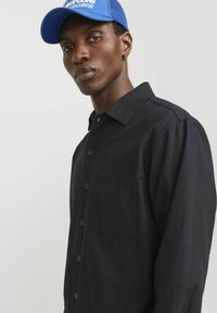 Chemise noire à boutons avec une poche poitrine et des coutures contrastantes, portée sous une casquette bleue avec un dos en maille et un logo.