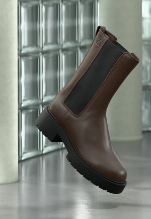 Brown leather ankle boot with black elastic side panel and chunky black sole against a glass block wall background.