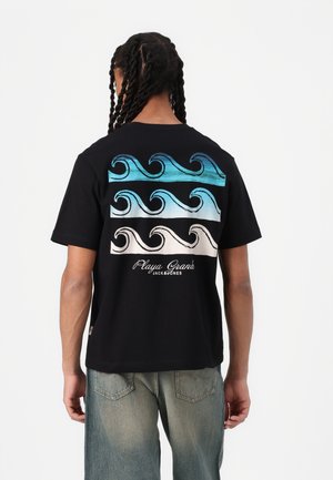 Personne aux cheveux tressés portant un t-shirt noir avec des motifs ondulés bleus et blancs et le texte "Playa Grande Jack & Jones", associé à un jean délavé.