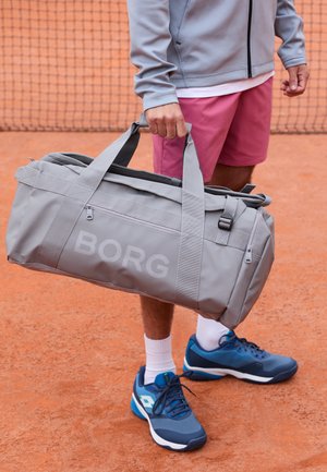 Pilkas sportinis krepšys su žodžiu "BORG" didelėmis raidėmis, su tvirtais rankenomis ir užtrauktuku uždarymu, padėtas ant teniso korto.