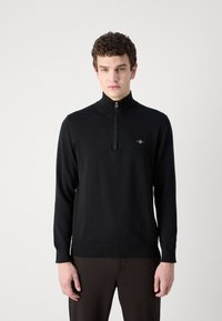 Crni džemper s patentnim zatvaračem i visokim ovratnikom, izrađen od pletenog materijala. Ima mali logo na prsima te rebraste rukave i rub.