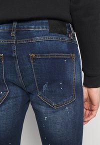 Tummansiniset farkut, joissa on slim fit -muotoilu. Niissä on kulutettuja kohtia ja valkoista maalisuihkua, ja ne on korostettu kontrastivärisellä keltaisella ompeleella.