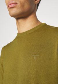 Gros plan d'une personne portant un pull en tricot vert olive avec un petit logo Barbour brodé sur la poitrine.