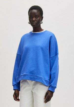 Sweatshirt bleu avec un col rond, tissu texturé, épaules tombantes et poignets côtelés. Associé à un pantalon de couleur claire.
