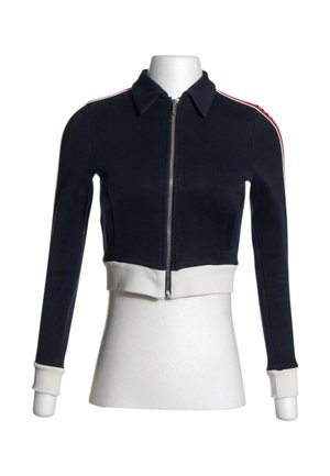 Veste courte bleu marine avec fermeture éclair frontale, poignets et ceinture blancs, et détails rayés rouge et blanc sur les épaules et les manches.