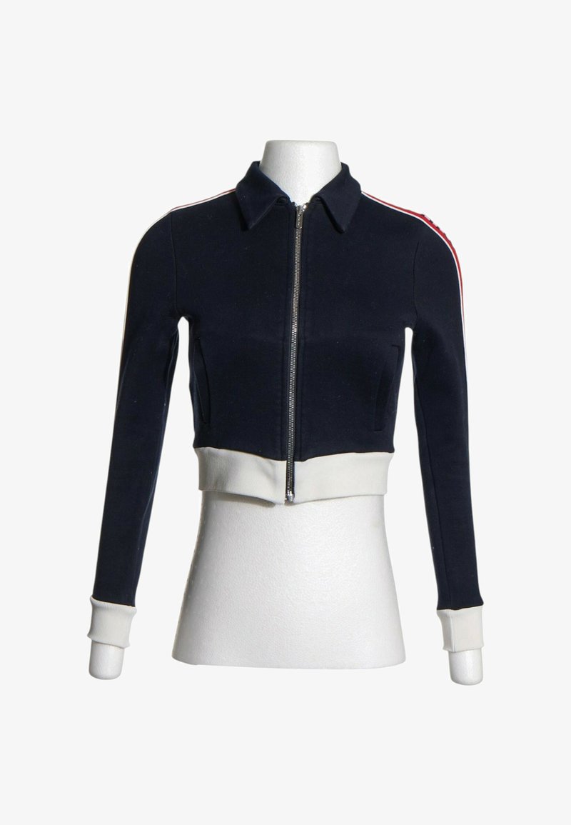 Veste courte bleu marine avec fermeture éclair frontale, poignets et ceinture blancs, et détails rayés rouge et blanc sur les épaules et les manches.