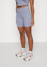 Damskie nogi w jasnofioletowych, wysokopodwyższonych szortach Nike, białych skarpetkach Nike i jasnoróżowych sneakersach Nike stojące na jednolitej podłodze.