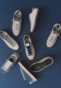 Sept baskets décontractées en blanc et bleu disposées sur une surface bleu foncé, certaines montrant le dessus et une montrant la semelle avec le logo.