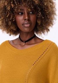 Mosterdgele trui met een losse pasvorm, wijd uitgesneden halslijn, een gestructureerd breipatroon en subtiele zijdetails. In combinatie met een zwarte choker met gouden accent.
