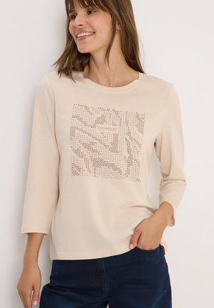 Chemise beige clair à manches longues avec un col rond, dotée d'un design texturé en beige plus foncé et d'un patch logo à l'avant.