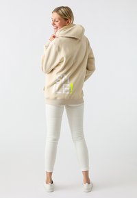 Beige hoodie with large back graphic reading "SMILE!" in white and neon green. Paired with white slim-fit pants and casual sneakers.