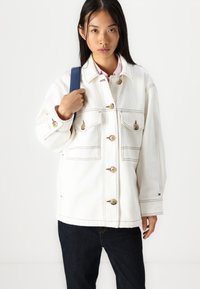 Jachetă din denim alb, cu patru buzunare frontale, nasturi mari și cusături vizibile. Asortată cu pantaloni din denim închis și o curea albastră.