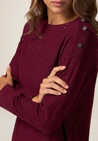 Pull en maille bordeaux avec un col et des poignets côtelés, orné de boutons décoratifs sur l'épaule et de paillettes dispersées.