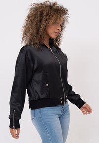 Zwarte satijnen bomberjack met geribde manchetten en zoom, voorzien van een ritsluiting en een zijzak, gedragen over lichtblauwe jeans.