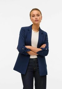 Navy blue blazer with rolled sleeves and button accents, paired with a white top and dark denim jeans. Smooth fabric, tailored design.