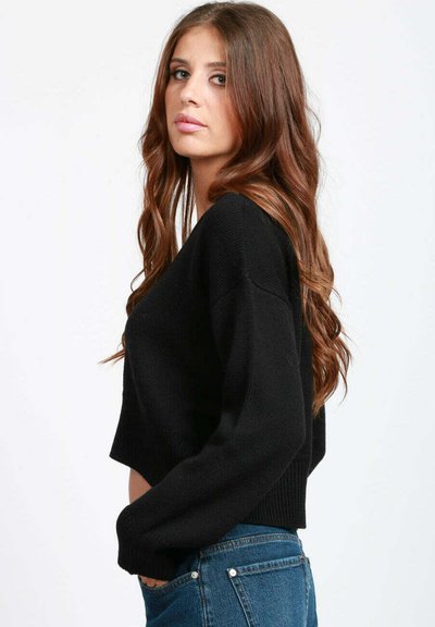 Giovane donna con lunghi capelli castani ondulati, che indossa un maglione nero e jeans blu, in profilo contro uno sfondo bianco semplice.