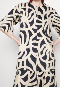 Top beige con motivo astratto nero, vestibilità comoda, maniche corte, scollo a V, abbinato a una gonna coordinata. La trama del tessuto appare liscia.