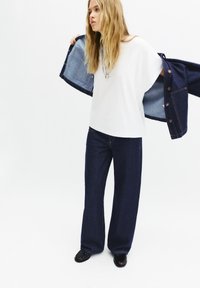 Cămașă albă cu mânecă lungă, pantaloni largi din denim închis și o jachetă din denim. Accesoriile includ un colier din argint. Materialul pare neted și lejer.