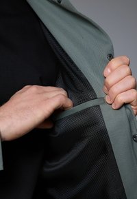 Camisa verde con una tela texturizada, que revela un forro de malla negra. Una mano está alcanzando el bolsillo interior con una correa en contraste.