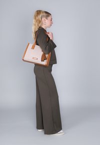 Vaalea nahkainen käsilaukku, jossa on teksturoitu vaaleanpunainen kangasrunko, nahkaiset yksityiskohdat ja tupsu. Mallilla on päällään oliivinvihreä tiukka asu ja lenkkarit.