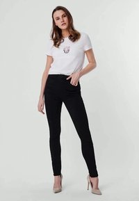 Černé skinny džíny v kombinaci s bílým tričkem s krátkým rukávem, které má růžovou květinovou grafiku. Modelka má na sobě béžové lodičky.