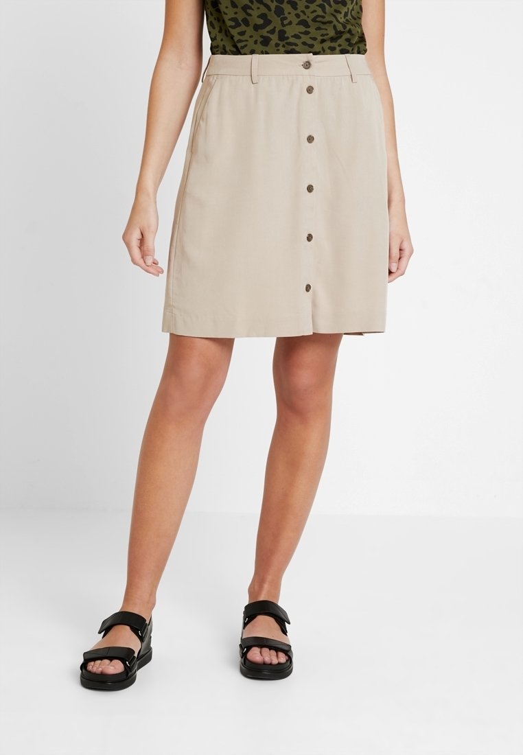 Beiger knielanger Rock mit Frontknopfverschluss, Seitentaschen und glatter Textur. Kombiniert mit schwarzen Sandalen vor hellem Hintergrund.