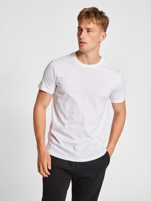 Junger Mann mit kurzen blonden Haaren, der ein schlichtes weißes T-Shirt und schwarze Hose trägt, steht mit einer Hand in der Tasche vor einfarbigem Hintergrund.
