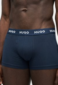 Torse masculin portant un boxer Hugo bleu marine avec le logo sur la ceinture élastique, sur un fond blanc uni.