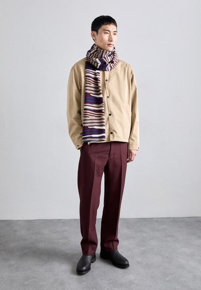 Giacca beige con bottoni neri, pantaloni borgogna scuri e una sciarpa a maglia a righe multicolore in crema, marrone, viola e blu. Calzature nere.
