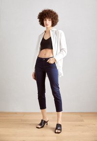 Biely košeľový top, čierny crop top, tmavomodré priliehavé džínsy a čierne sandále na vysokých podpätkoch. Podlaha je svetlé drevo. Pozadie je neutrálne.
